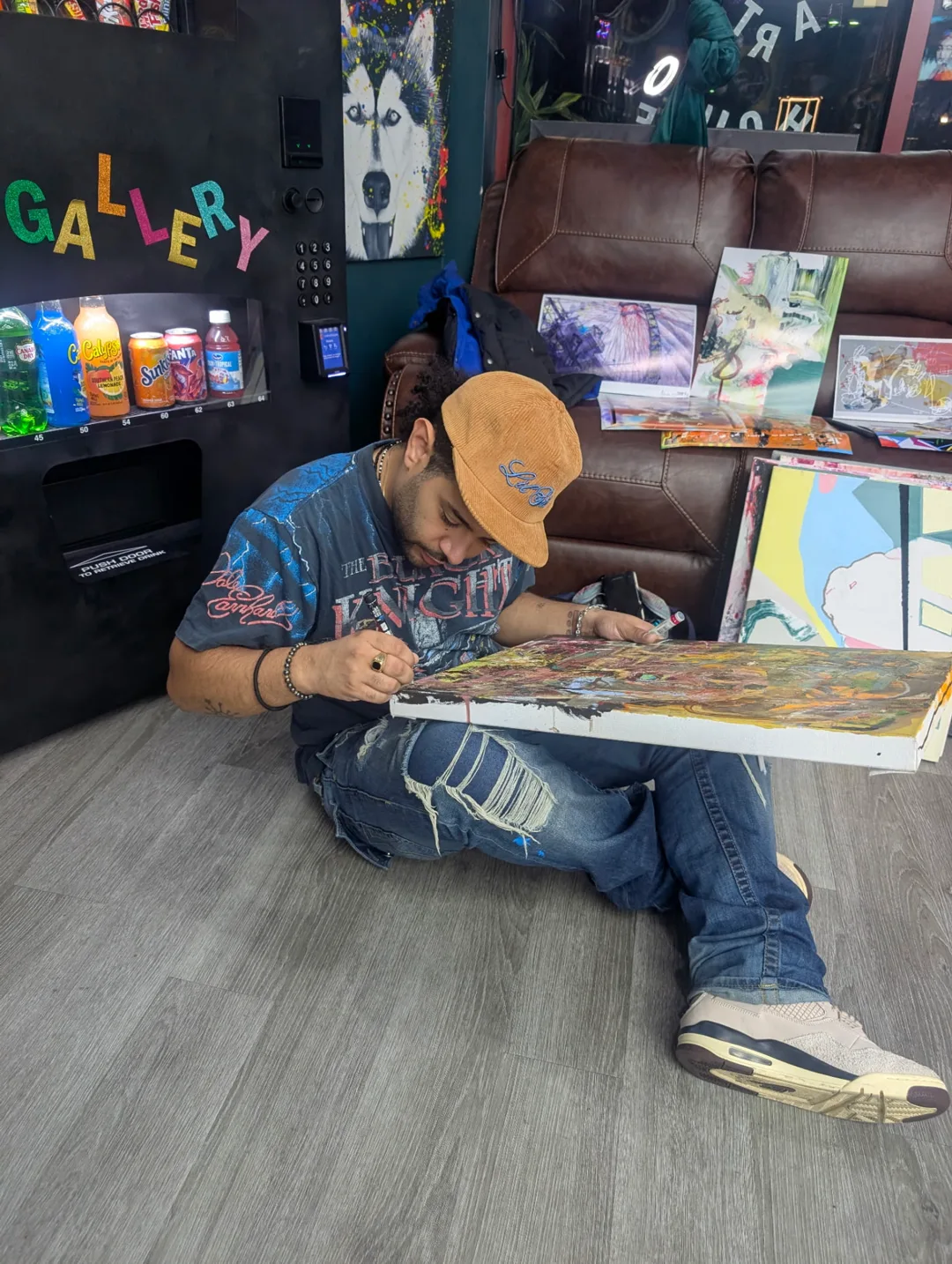 An artist at work on a canvas on the gallery floor, surrounded by finished paintings